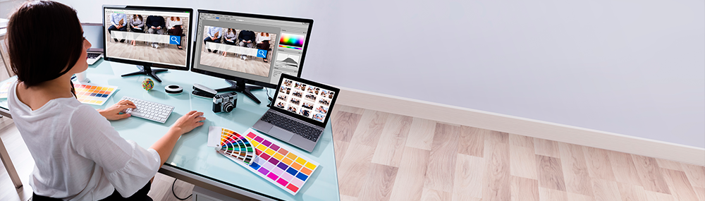 Kobieta przy komputerze, dwa monitory i laptop oraz aparat fotograficzny i wzorniki barw